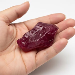 Puede incluir: Una gema de rubí en bruto, sin cortar, de color rojo intenso. El rubí se sostiene en una mano abierta, mostrando su forma irregular y textura natural. La superficie de la gema refleja la luz, resaltando su rico color y estructura cristalina.