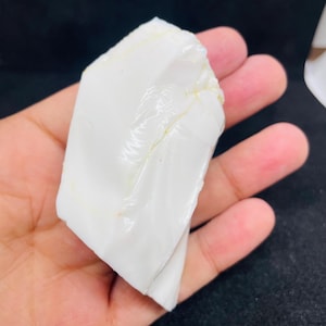 May include: A large, irregularly shaped, opaque white stone with a glossy surface. The stone is held in a hand against a dark background. The stone appears to be a natural mineral or a piece of raw material.