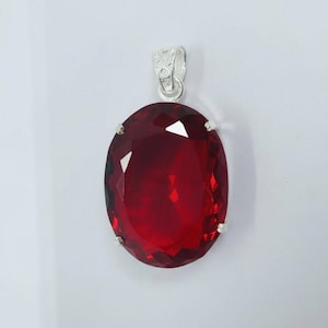 Pode incluir: Um pendente de prata com uma grande gema vermelha escura, facetada e de forma oval. A gema é engastada numa moldura de prata e tem uma argola de prata. O pendente é mostrado sobre um fundo branco. Mede aproximadamente 3 cm x 2 cm.