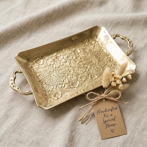 May include: A rectangular gold-toned metal tray with ornate floral embossing and two side handles. A small bouquet of dried flowers is tied to the tray with twine, accompanied by a brown paper tag that reads Handcrafted For a Special Home.