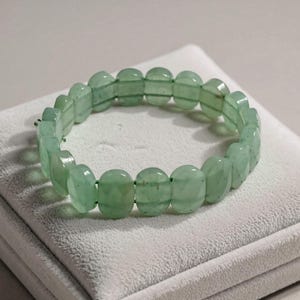May include: A light green beaded bracelet. The bracelet is made of oval-shaped, faceted beads strung together. The beads have a translucent quality, and the bracelet is resting on a white surface.