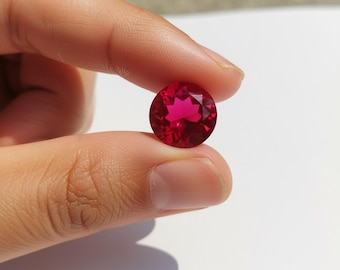 May include: A faceted, round, deep red gemstone. The gem is held between two fingers against a white background. The gem's cut creates multiple facets that reflect light, giving it a brilliant appearance. The color is a rich, saturated red.