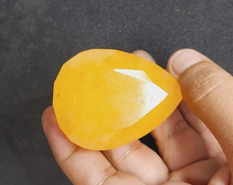 May include: A faceted, teardrop-shaped gemstone in a vibrant yellow hue. The gem has multiple facets that reflect light, creating a sparkling effect. The stone is held in a hand against a dark background.
