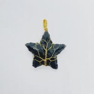 Puede incluir: Un colgante negro en forma de estrella con un envoltorio de alambre dorado. El colgante está hecho de un material oscuro, posiblemente obsidiana. El alambre está envuelto alrededor de los bordes de la estrella y forma un lazo en la parte superior para colgar.