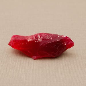 May include: A vibrant, irregularly shaped, deep red gemstone. The polished surface reflects light, showcasing the stone's rich color and natural texture. The gemstone is set against a neutral background.