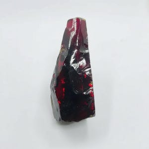 Puede incluir: Una gema en bruto, de color rojo oscuro, con una superficie brillante y reflectante. La piedra tiene forma triangular y parece ser un espécimen mineral natural. El color varía del burdeos oscuro al casi negro en algunas zonas.
