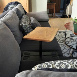 Könnte beinhalten: Ein Beistelltisch aus Holz mit quadratischer Platte und schwarzem Metallsockel. Der Tisch besteht aus hellbraunem Holz und wird von einer einzelnen schwarzen Metallsäule getragen. Der Tisch ist für die Platzierung neben einem Sofa konzipiert.