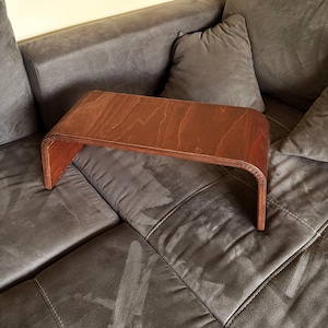 Peut inclure: Un plateau de bras de canapé en bois, de couleur marron chaud. Le plateau a un dessus rectangulaire avec des coins arrondis et une base incurvée, conçu pour reposer sur un accoudoir de canapé. Le grain du bois est visible.