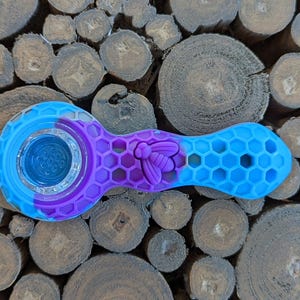 May include: A blue, purple, and clear glass pipe with a bee design. The pipe has a honeycomb pattern on the stem and a clear glass bowl. The pipe is made of silicone and glass.