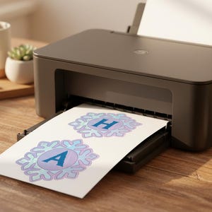 Op de afbeelding: Een donkergrijze printer print twee sneeuwvlokvormige ontwerpen met de letters "A" en "H" in blauw. De ontwerpen staan op wit papier. De printer staat op een houten oppervlak, met een kleine vetplant en potloden op de achtergrond.