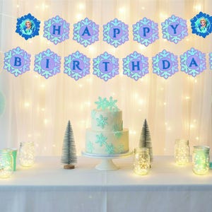 Op de afbeelding: Een verjaardagsfeestdecoratie met een taart met twee lagen, ballonnen en een banner met de tekst "HAPPY BIRTHDAY". De taart is versierd met sneeuwvlokken. Het kleurenschema is lichtblauw en wit.