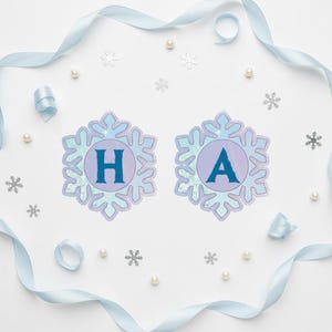 Op de afbeelding: Een feestelijke banner met de letters "HA" in blauwe letters, elk in een sneeuwvlokvormig ontwerp. De banner is omgeven door lichtblauw lint, imitatieparels en sneeuwvlokconfetti op een witte achtergrond.