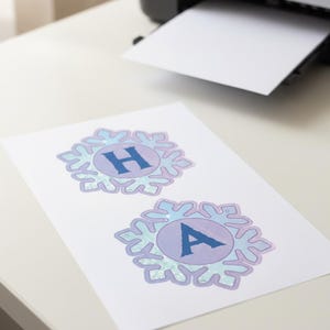 Op de afbeelding: Een vel papier met twee sneeuwvlokvormige ontwerpen, elk met een letter. Het bovenste ontwerp heeft de letter "H" en het onderste ontwerp de letter "A". De ontwerpen zijn in blauwe en paarse tinten. Een printer is op de achtergrond zichtbaar.
