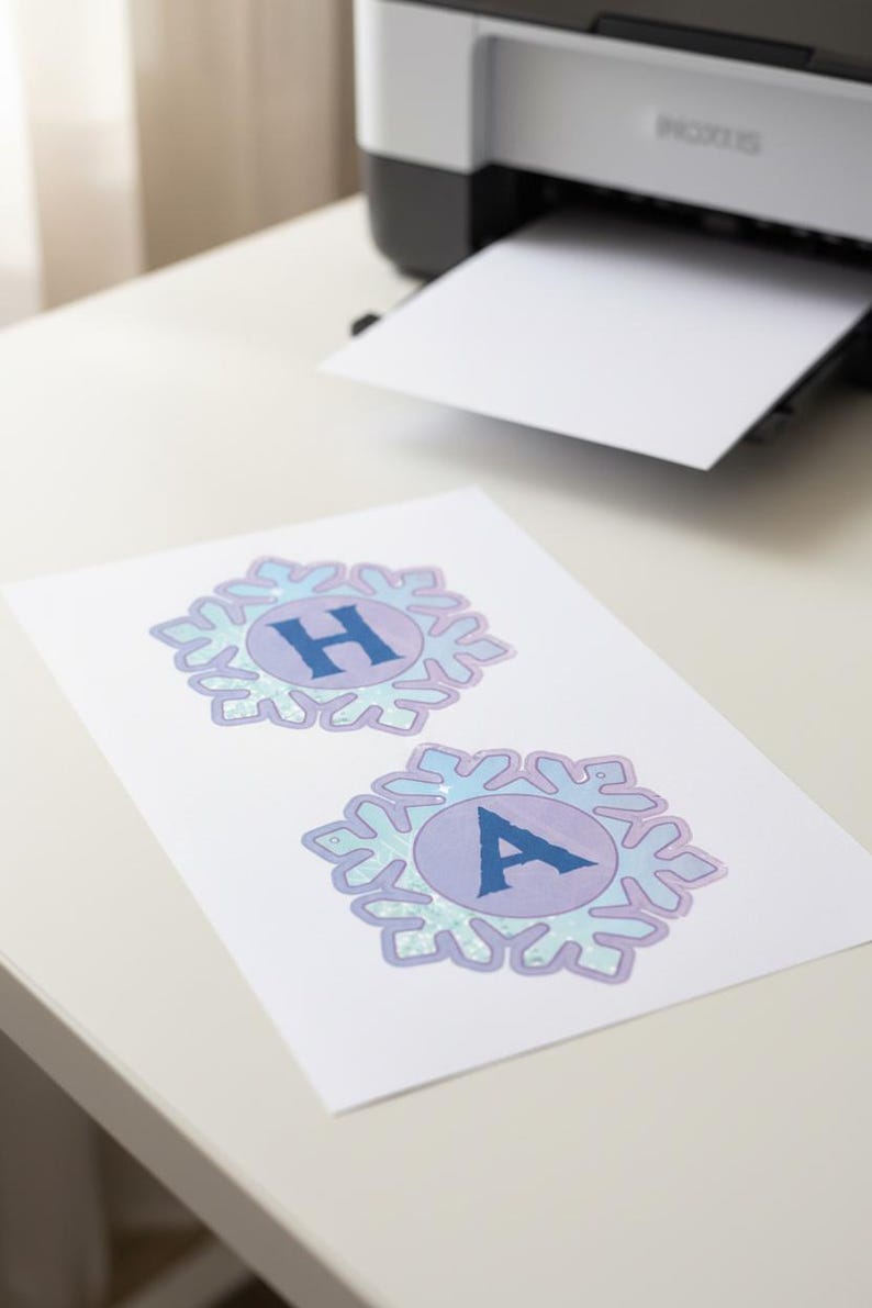 Op de afbeelding: Een vel papier met twee sneeuwvlokvormige ontwerpen, elk met een letter. Het bovenste ontwerp heeft de letter "H" en het onderste ontwerp de letter "A". De ontwerpen zijn in blauwe en paarse tinten. Een printer is op de achtergrond zichtbaar.
