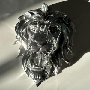 May include: A polished silver-coloured wall sculpture depicting a lion head with a full mane and an open mouth showing teeth. The metallic surface reflects light, creating high-contrast highlights and shadows across the detailed facial features.