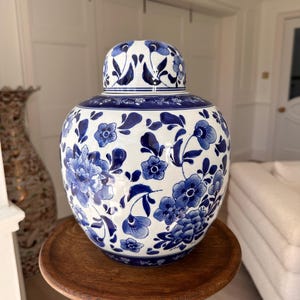 May include: A blue and white ceramic ginger jar with a lid, decorated with a floral pattern. The jar is round and rests on a small, round wooden table. The floral design is in blue against a white background.