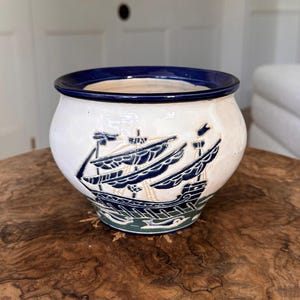 Puede incluir: Una maceta de cerámica blanca con un borde azul marino y una ilustración detallada de un velero. El barco está representado con velas azul marino y detalles dorados, sobre un fondo blanco. La base de la maceta tiene un borde verde.