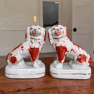 Puede incluir: Dos figuras de perro de cerámica. Los perros son blancos con manchas rojas y collares marrones. Se asientan sobre bases rectangulares con ribetes dorados. Las figuras están sobre una superficie de madera.