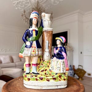 Op de afbeelding: Een vintage keramieken beeldje met twee figuren in traditionele Schotse klederdracht. De figuren dragen blauwe jassen, kilts en kleurrijke hoeden. Het beeldje heeft een bruine zuil met een witte kan en een muziekinstrument. De basis is versierd met geel en groen gebladerte.