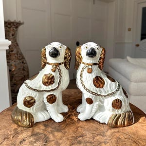 Puede incluir: Dos figuras de perros de cerámica. Los perros son blancos con manchas marrones y detalles dorados en las orejas, patas y colas. Cada perro tiene un collar dorado con una cadena. Sobre una mesa de madera.