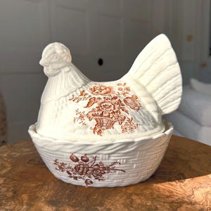 Könnte beinhalten: Eine weiße Keramik-Henne-förmige Schale mit einem Korbgeflecht-Boden und einem braunen Blumendesign. Der Kopf und der Schwanz der Henne sind erhöht, und der Deckel hat ein Blumengestaltung. Ein dekorativer Küchenartikel.