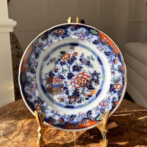 May include: A decorative plate featuring a floral pattern in blue, orange, and white. The plate is presented on a gold-coloured stand. The rim of the plate has a dark blue border with orange detailing.