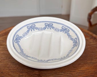 May include: Oval white ceramic soap dish with a blue floral and bow design around the rim. The dish has three raised bars in the center for soap placement. The dish is sitting on a wooden surface.