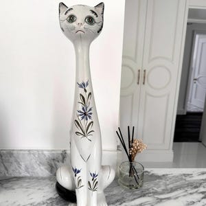 May include: A white ceramic cat figurine with a long neck and seated pose. The cat has green eyes, black markings, and is decorated with blue floral accents. The figurine is displayed on a marble surface.