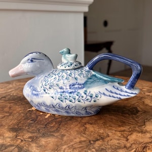 May include: A ceramic duck teapot with a blue and white floral pattern. The teapot has a duck-shaped body, a blue handle, and a small duck figurine on the lid. The beak is pink.