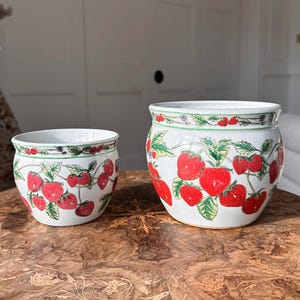 May include: Two white ceramic pots adorned with a cheerful cherry pattern. The pots display a repeating design of vivid red cherries and green leaves on a white base. The rims are decorated with a green border and additional cherry and floral accents.