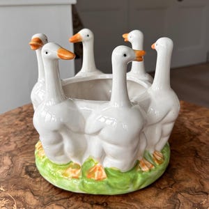 May include: A white ceramic planter with six geese around the rim. The geese have orange beaks and feet, and are set on a green base. The planter is a decorative item for home decor.