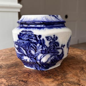 Könnte beinhalten: Ein weiß-blaues Keramikgefäß mit Deckel. Das Gefäß hat ein Blumendesign in Blautönen. Das Gefäß steht auf einer Holzoberfläche. Das Gefäß hat eine sechseckige Form.