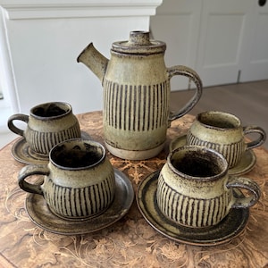 May include: A ceramic tea set with a teapot and four cups and saucers. The set has a speckled, earthy green glaze with vertical brown stripes. The cups and teapot have rounded shapes and curved handles.