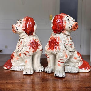 Puede incluir: Un par de figuras de perros de cerámica. Los perros sentados tienen cuerpos blancos y marcas rojas en las orejas y la espalda. Tienen collares de cadena y están colocados sobre una superficie de madera.