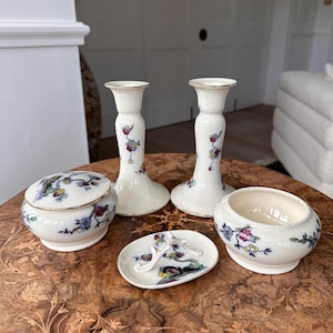Puede incluir: Un conjunto de artículos de porcelana vintage, que incluye dos candelabros, una caja de baratijas con tapa, un plato pequeño y un cuenco redondo. Cada pieza es blanca con ribetes dorados y diseños florales en azul, rosa y amarillo.