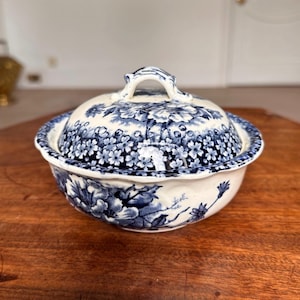 May include: A white and blue ceramic serving bowl with a lid. The bowl and lid feature a floral pattern in shades of blue. The bowl is round with a scalloped edge and a handle on the lid.
