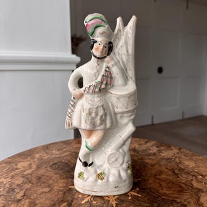Puede incluir: Figura de cerámica vintage de un baterista escocés con vestimenta tradicional. La figura es predominantemente blanca con acentos rojos, verdes y negros. El baterista lleva un sombrero, un kilt y sostiene un tambor. La figura mide aproximadamente 20 cm de altura.
