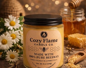 May include: A jar candle with a black lid and a label that reads "Cozy Flame Candle Co." The label also states "Made with 100% Pure Beeswax" and "Net Wt 8 oz (226g)." The candle is surrounded by daisies, honey, and a honeycomb.