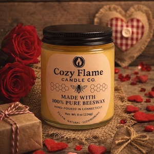 Puede incluir: Una vela en tarro con tapa negra y una etiqueta que dice "Cozy Flame Candle Co.". La etiqueta también indica "Hecho con 100% cera de abeja pura". La vela está rodeada de rosas rojas, decoraciones en forma de corazón y una pequeña caja de regalo.