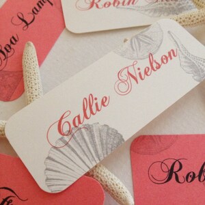 Peut inclure: Cartes de place roses et blanches avec un motif de coquillage et des noms écrits en calligraphie noire. Les noms incluent Callie & Nielson, Robin, Boa Lampton, Frazer et Rob.