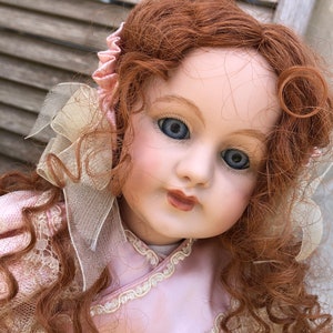 Peut inclure: Une poupée en porcelaine aux longs cheveux roux bouclés, portant une robe rose avec des bordures en dentelle et un nœud blanc.