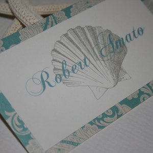 May include: A white place card with a teal border and a seashell design. The name "Robert Imato" is written in cursive on the card.