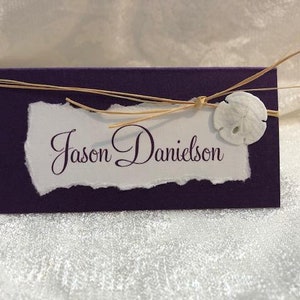 Peut inclure: Carte de place violette avec une étiquette en papier blanc qui dit "Jason Danielson". L'étiquette est attachée avec un ruban de raphia naturel et un coquillage blanc.