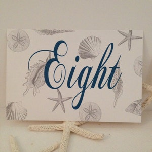 May include: A white card with a navy blue seashell design and the word "Eight" written in a decorative script font. The card is surrounded by white starfish.