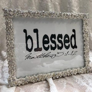 Peut inclure: Un cadre photo argenté avec une bordure en strass. Le cadre contient un passe-partout blanc avec le mot "blessed" en lettres noires et le texte "Matthew 5:1-12" en écriture cursive noire.