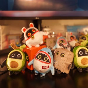 Op de afbeelding: Een verzameling pluche sleutelhangers met verschillende ontwerpen, waaronder een haai, avocado en andere stripfiguren. De haai sleutelhanger heeft het woord "SHARK" erop. De sleutelhangers zijn in verschillende kleuren, waaronder blauw, groen en wit.