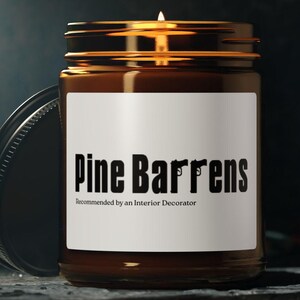 Peut inclure: Une bougie allumée dans un bocal en verre marron avec un couvercle noir. L'étiquette indique "Pine Barrens" avec un motif de pistolet. Le texte "Recommandé par un décorateur d'intérieur" est en dessous. La bougie est sur une surface sombre.
