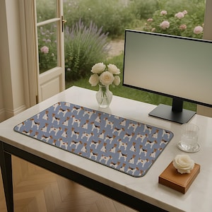 May include: A rectangular desk mat featuring a repeating pattern of terrier dogs in various poses on a light blue background. The mat is on a white desk with a computer monitor, a vase of white roses, and a glass of water.