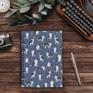 May include: Spiral-bound notebook with a blue cover featuring a repeating pattern of black and white Border Collie dog illustrations. The notebook is open, revealing the patterned cover. The image is set on a wooden surface with a pen, camera, and clock.