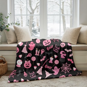 Könnte beinhalten: Schwarze Halloween-Decke mit rosa Illustrationen. Das Design umfasst Geister, Kürbisse, Fledermäuse, Spinnennetze, Süßigkeiten und die Wörter "Boo!" und "Trick or Treat". Die Decke liegt auf einer Fensterbank.